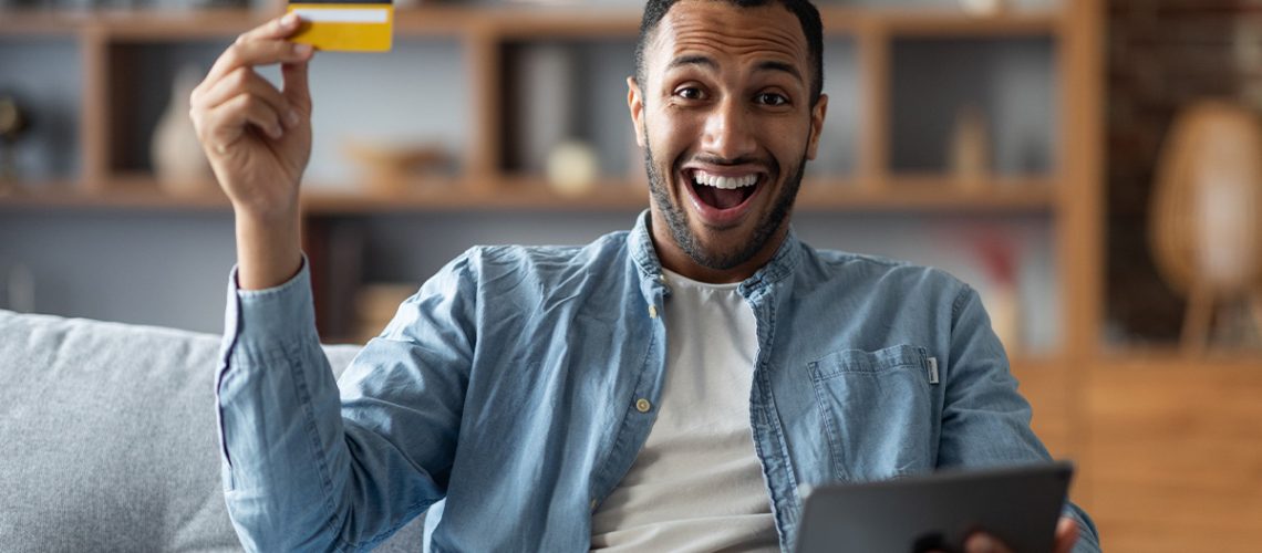 A man is sitting on a couch in his home holding a credit card and tablet and smiling