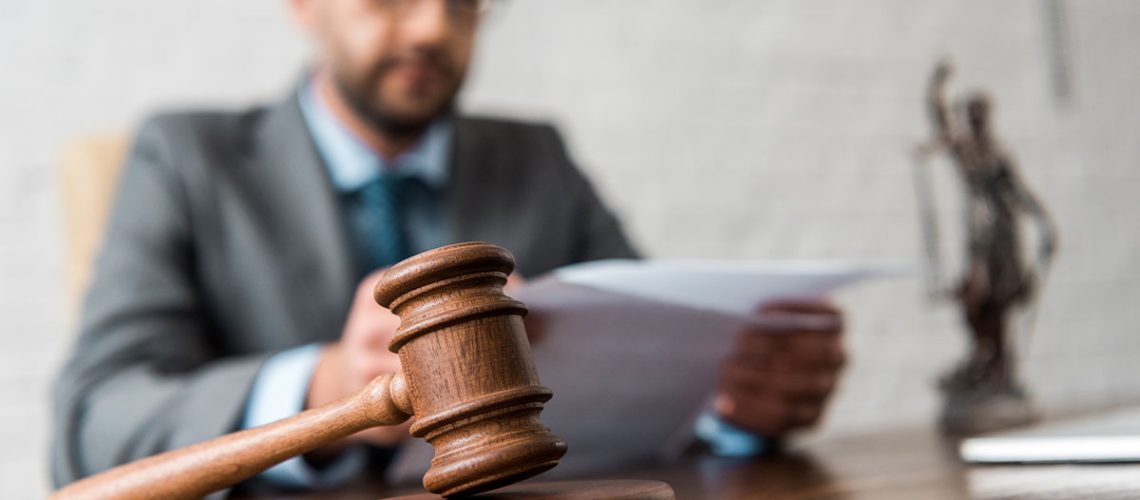 A bearded man with black glasses, grey blazer, white shirt and blue tie is sitting at a wooden desk reviewing papers with a gavel and statue of justice next to him.