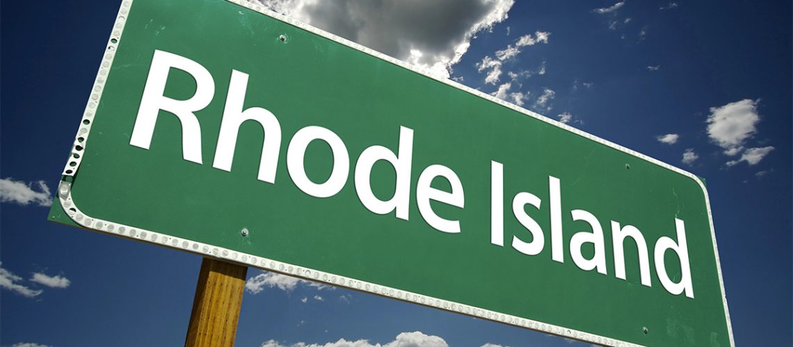 A green and white road sign that reads "Rhode Island" with a blue sky and clouds behind it