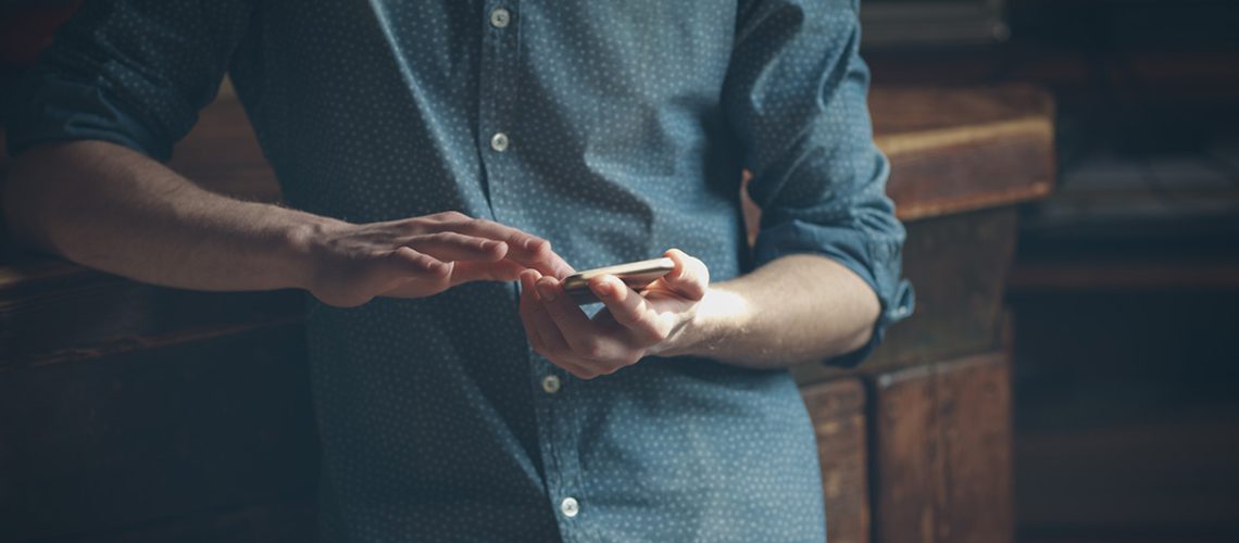 Man with a light blue button down shirt leaning on a wooden tabletop holding his phone and texting