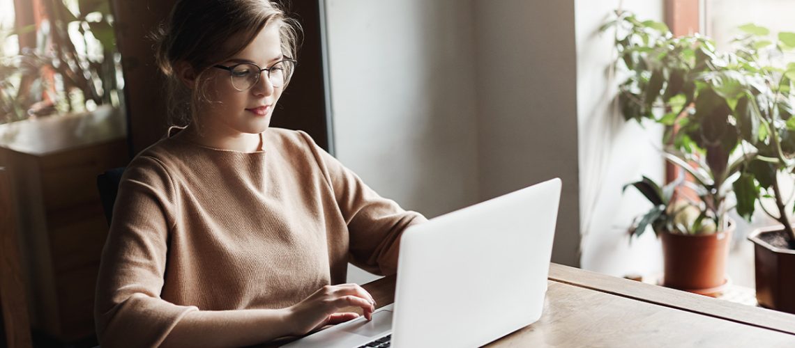 A woman with glasses sitting at a table in her home is working on her laptop