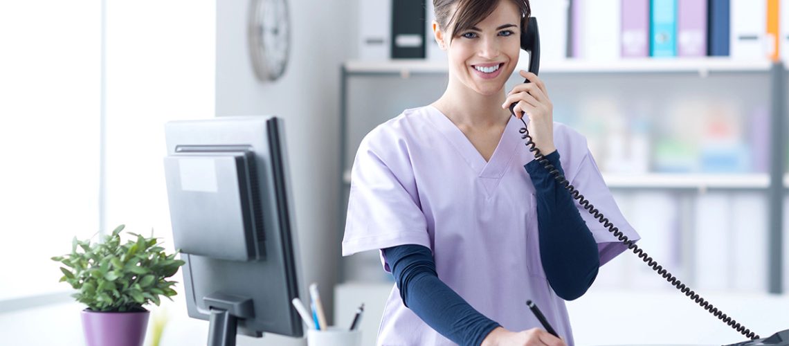 A medical receptionist is smiling on the phone and writing notes on a clipboard