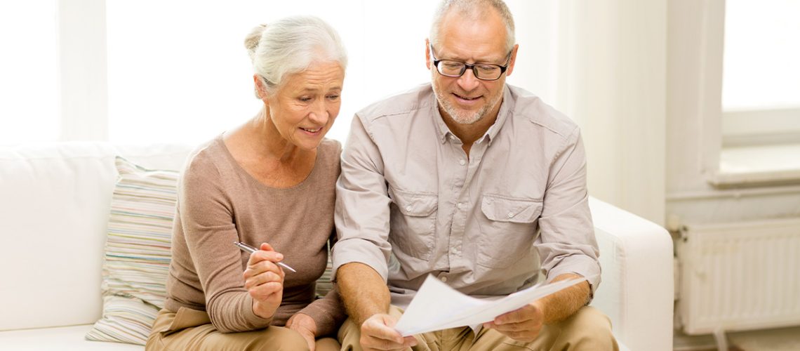 A senior couple wearing neutral colors are sitting on a couch and reviewing paperwork