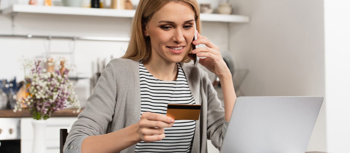 A woman is sitting in her home at the kitchen table while on the phone looking at her credit card and her laptop in front of her on the table