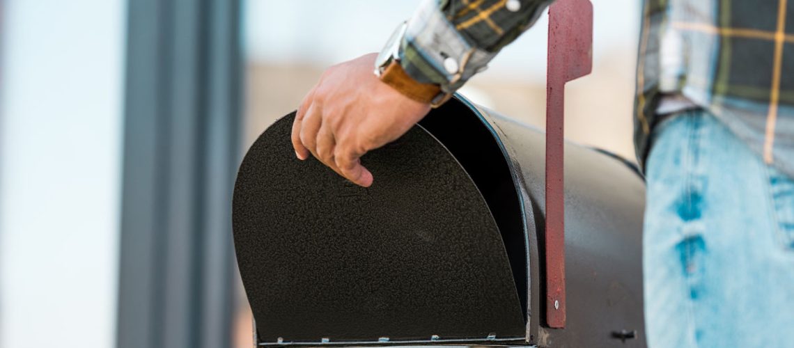 A male wearing a flannel shirt and light blue jeans closes a black mailbox to send a payment