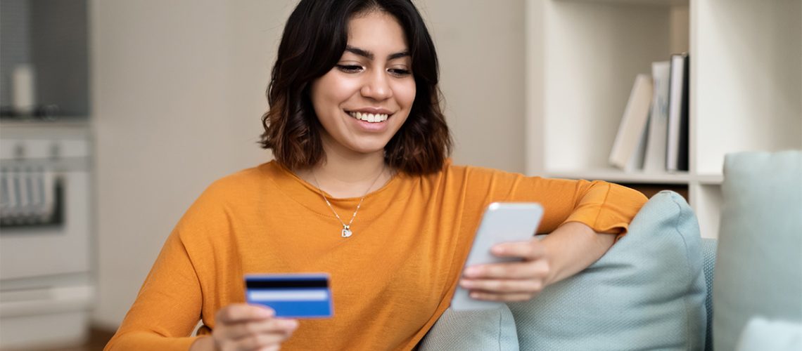 Woman with short dark hair wearing a mustard yellow top is sitting on a couch in her home making a credit card payment on her phone